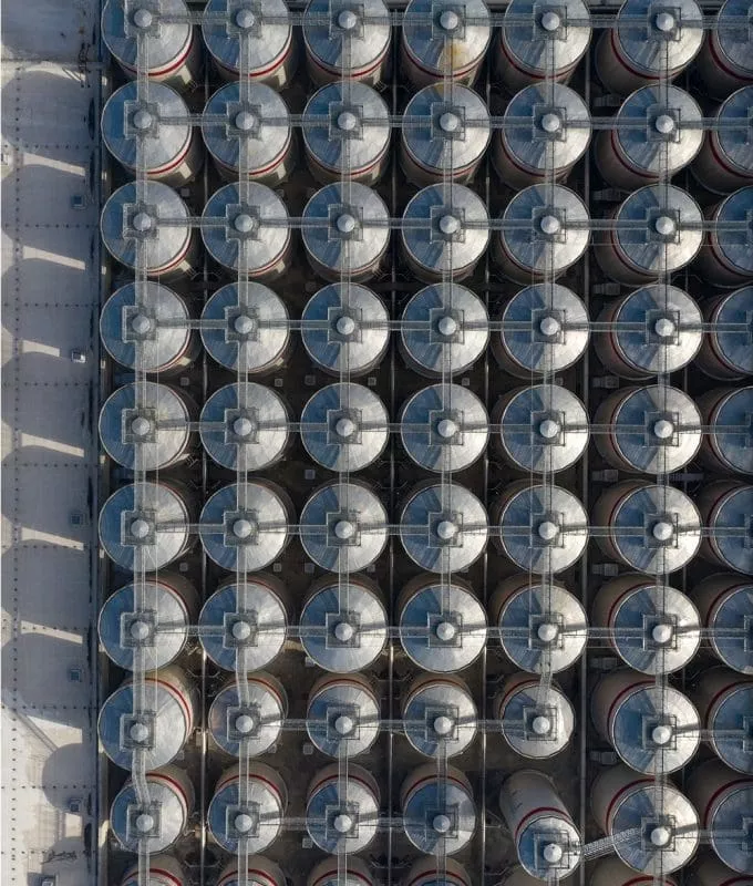 Aerial view of an industrial facility with rows of large cylindrical storage tanks arranged in a grid pattern next to a flat-roofed building.