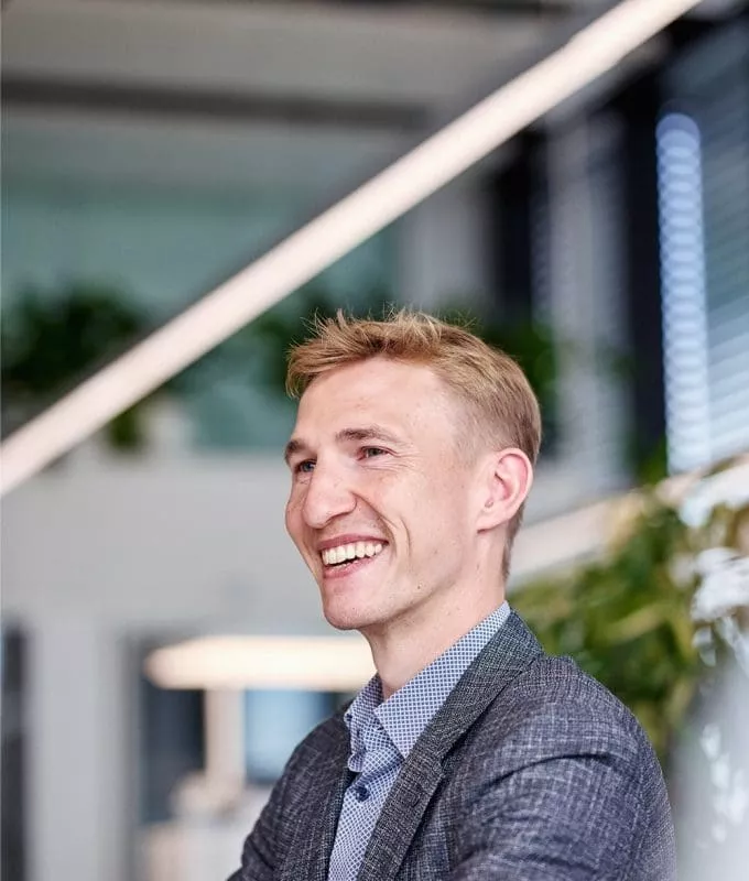 Smiling man in a checkered blazer and blue shirt in a bright, modern office with plants and large windows in the background.