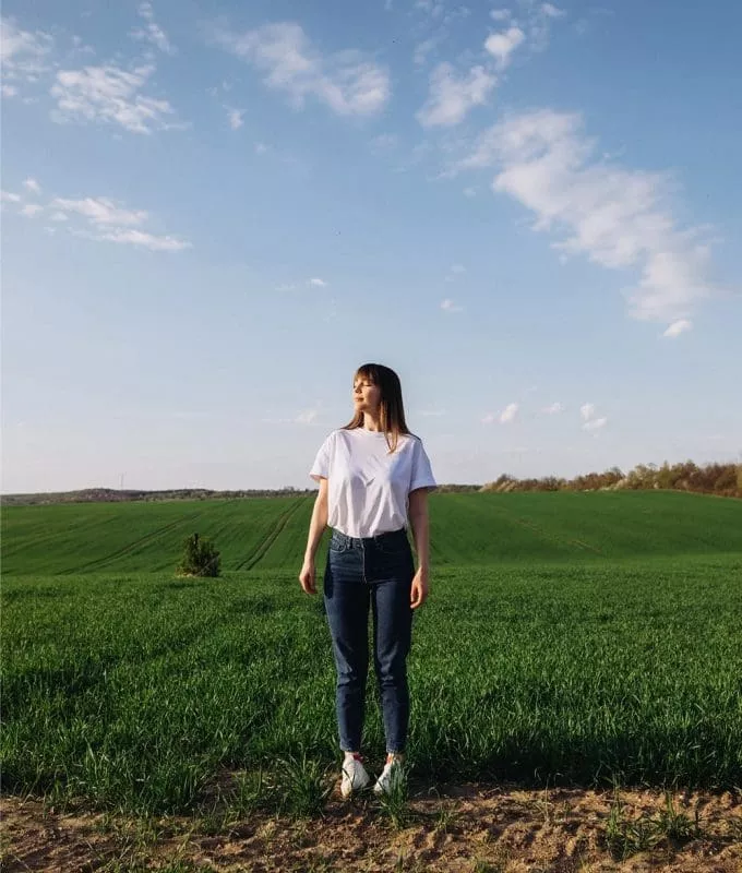 Woman standing in a green field under a blue sky with scattered clouds, wearing a white T-shirt and jeans, looking off into the distance.