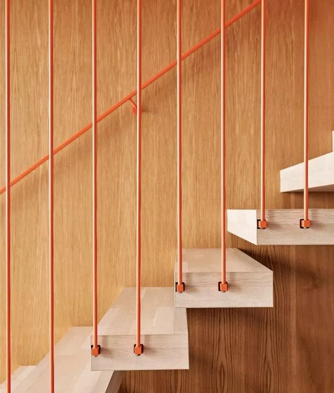 Modern staircase with light wooden steps and vertical orange metal railings set against a wooden panel wall.